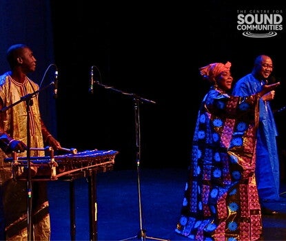 From left to right: Fodé Lassana Diabaté, a Black man wearing a light-colored tunic and dark red shirt, playing the balafoon (a struck instrument similar to a xylophone); Hawa Kassé Mady Diabaté, a Black woman wearing a patterened cloth dress and red headscarf, singing with arms outstretched, and Chérif Keita, a Black man in a blue robe, smiling.