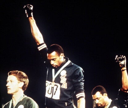 Medal podium at the 1968 Olympic Games. Tommie Smith (the gold medalist, center), a Black man, is wearing a dark blue Team USA jacket (with red and white trim), the gold medal around his neck, and his right hand in a Black Power salute with a black glove on his clenched right fist. John Carlos (bronze medalist, right), a Black man, is also wearing a Team USA uniform (with an unzipped jacket) and mirrors the salute with his left hand. Peter Norman (left), a white man, is wearing a green jacket.