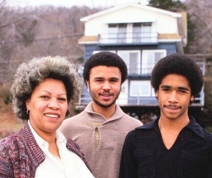 Toni Morrison, a Black woman wearing a multicolored sweater, white blouse, and black pants, next to her son Harold (wearing a brown sweater and khaki pants) and Slade (wearing a black shirt and red pants), in front of a multi-story house and trees.