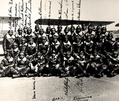 Several Black men in three rows (sitting, kneeling, standing), all in dark flight suits, boots, and goggled helmets, in front of biplanes. Some men's signatures are next to their photograph.