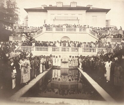 Black and white photograph of a large group of people, mostly women, posed around the exterior of a large Italianate style mansion. The figures are arranged on the building's multi-level balconies, stairs, and around a rectangular pool on the ground level. The photograph is mounted on a piece of gray cardboard that has sustained loss at the top right and bottom right corners. In the bottom right corner is black script that is partially missing.