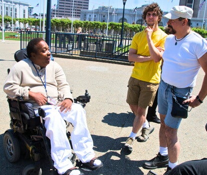 A Black man wearing a tan sweater and white pants in a power wheelchair, talking to a white man in a white hat, white shirt, jean shorts, white socks, and black boots, as another white man in a yellow shirt, khaki shorts, white socks, and sneakers looks on.