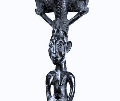 A wood sculpture, painted black, in the form of a standing female figure with arms at sides apart from torso, hands on hips, and feet and legs apart; on her head there is a double-ax motif.