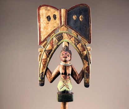 Wooden staff in the form of a kneeling female figure holding a stylized snake in an inverted U over her head, topped by a pair of opposing triangles in a bowtie or double-ax form. Female figure has white beads around her neck and staff is pigmented in red, blue, and white.