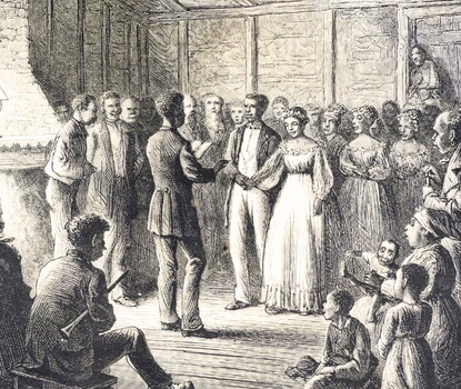 A wedding near a fireplace. The groom is a Black man wearing a dark coat, white pants, and a white shirt. The bride is a Black woman wearing a white dress. Groomsmen and bridesmaids are behind the couple, and the officiant (wearing a dark coat and pants and holding a book) is faced in the couple's direction. Behind the officiant are more witnesses, a bassist, and a clarinetist (the latter musicians sitting).