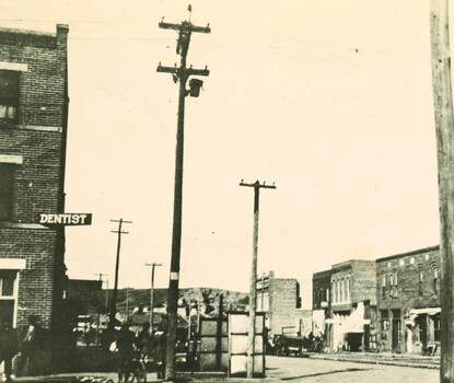 A black-and-white photograph of the Greenwood district in Tulsa, Oklahoma, depicting two- and three-story brick buildings, storefronts, telephone poles, and a sign for a dentist's office. People are visible on the sidewalk.