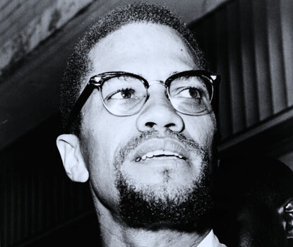 A black-and-white photograph of a goateed Malcolm X wearing horn-rimmed glasses and a striped suit, white shirt, and dark tie, looking toward his left.