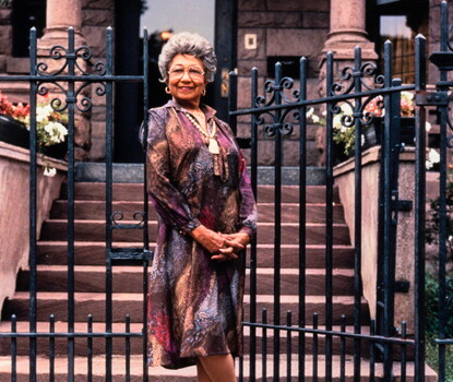 Etta Moten Barnett, a Black woman wearing a multicolored dress and white necklace, standing next to the metal gates in front of her house.