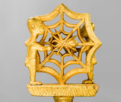 A gold staff with two human figures on either side of a spider web.