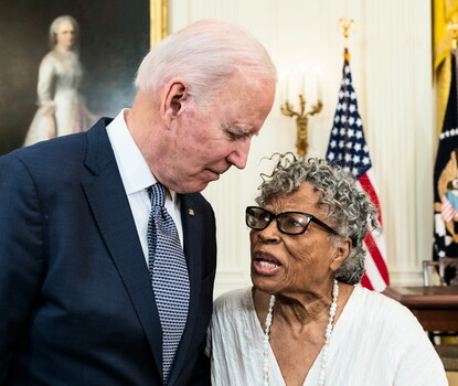 Joe Biden, wearing a blue suit, blue dotted tie, and flag lapel pin, standing next to and listening to Opal Lee, a bespectacled Black woman wearing a white shirt and necklace.