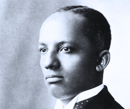 A black-and-white photograph of Carter G. Woodson, a Black man, wearing a dark jacket, white shirt, and polka-dotted tie