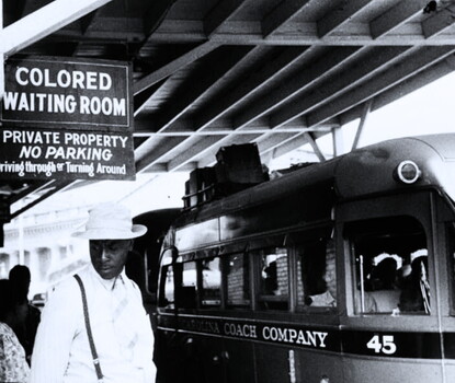 A black-and-white photograph of a Black man wearing a light-colored shirt, tie, and hat, and dark suspenders, under a sign reading "Colored Waiting Room."