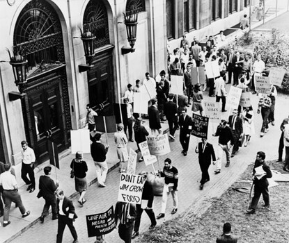 A black-and-white photograph of a crowd picketing with signs outside a building, photographed from above.