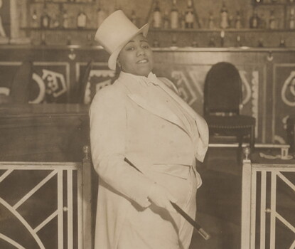 A sepia photograph of entertainer Gladys Bentley in three-quarter profile, wearing a white tuxedo and top hat, holding a cane under her right arm.