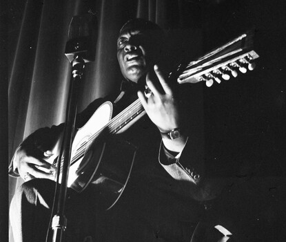 Lead Belly, a Black musician, playing a guitar and singing into a microphone in front of a dark background.