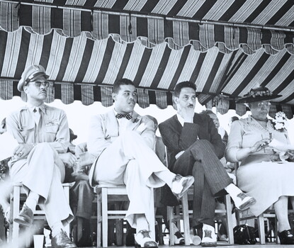A black-and-white photograph of Benjamin O. Davis, Jr. (a Black soldier in uniform and sunglasses), Joe Louis (a Black man wearing a light colored suit and a dark bowtie), Thurgood Marshall (a Black man wearing a dark suit and white shirt, his head resting on his crooked right arm), and Daisy Lampkin (a Black woman wearing a light colored dress and dark hat) seated outside under an open-air tent.
