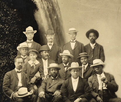 A black-and-white image of a photograph of twelve Black men in three rows of four (and a child on one man's knee, second row left), all wearing dark suits and hats, superimposed on a photograph of Niagara Falls.