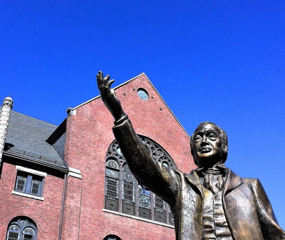 A bronze statue of Richard Allen, a Black man, wearing a jacket, vest, and ruffled collar shirt, with his right arm outstretched. Behind the statue is a brick church building to the right of a stone church building.