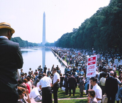 A color photograph of a crowd gathering around the reflecting pool in Washington, the Washington Monument, and the Capitol rotunda in the distance. In the foreground, a White man holds a red-and-white sign reading "We march for jobs for all now!"