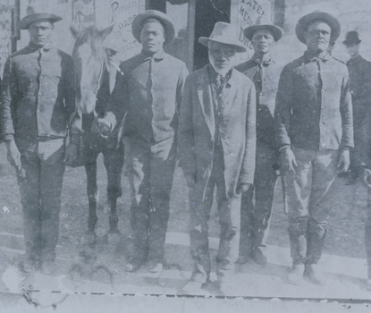 A black-and-white photograph of seven men wearing wide-brimmed hats. To the right of the man third from left is a horse.