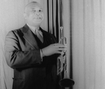 A black-and-white photograph of W. C. Handy, a Black man wearing a dark suit, white shirt, and light tie, holding a trumpet.