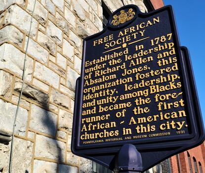 A color photograph of a blue historical marker with yellow text for the Free African Society, in front of a gray stone building.