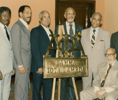 A color photograph of six standing (and one seated) Black men in suits and ties, in front of a metal sign on a wooden easel reading "Alpha Phi Alpha [in Greek letters]: Gamma Iota Lambda."