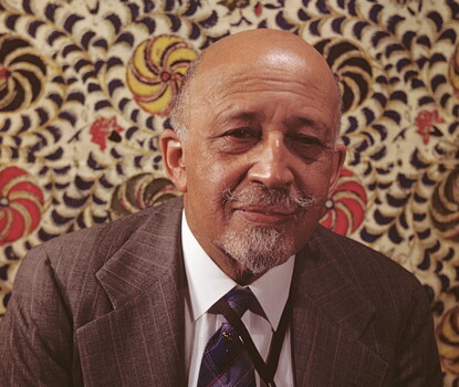 A color photograph of W. E. B. Du Bois, wearing a brown pinstriped suit, white shirt, and blue-and-gray checked tie, against a background of multicolored spirals.