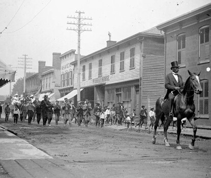 A black-and-white photograph of a Black man wearing a dark top hat and suit riding a horse leading a procession including a band of musicians down a dirt road