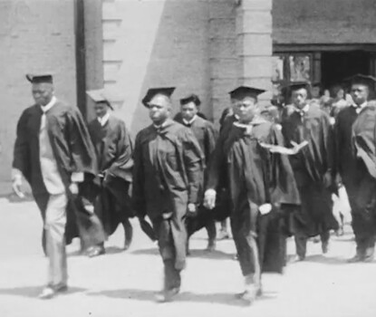 A black-and-white still from a film of a graduation procession of Black men in black caps and gowns with Black women in white dresses behind them.