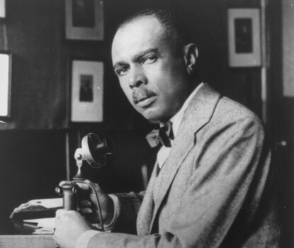 A black-and-white photograph of James Weldon Johnson, a Black man wearing a light colored blazer, white shirt, and dark bow tie, seated at a desk, turning left toward the camera. He is holding a telephone in his left hand.