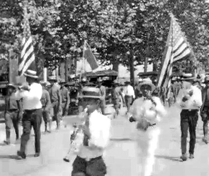 A still from a black-and-white film showing Black men and women, many in straw-boater hats with dark hatbands, marching in a parade. In the foreground is a clarinetist, followed by two American flag bearers marching at the head of the parade.