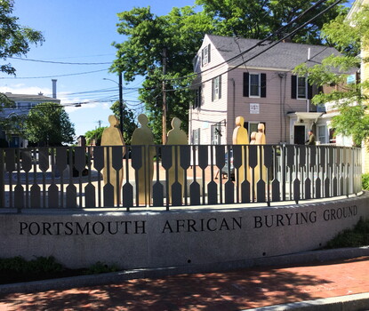 A color photograph of six golden silhouette statues in a semicircle, behind a fence, with a pedestal below reading "Portsmouth African Burial Ground."