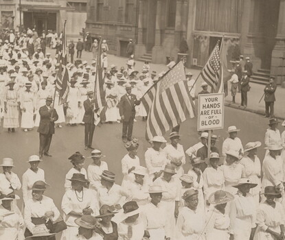 A black-and-white photograph of a parade; Black women, most wearing white dresses and hats, march in formation in rows of about twenty. Between two groups of women there is a row of four Black men in dark suits, ties, and hats, holding up large American flags. In the first group of women, one woman holds up a sign reading, "YOUR HANDS ARE FULL OF BLOOD."