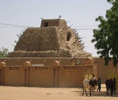 The Tomb of Askia, a mud building covered in spikes, surrounded by two sets of walls. On the other side of the outer wall, cattle walk behind three children.