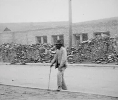 A black-and-white photograph of a Black man wearing a light-colored suit and dark bowler hat walking with a cane in his right hand; in the background is rubble of buildings destroyed in the Tulsa Race Massacre. 