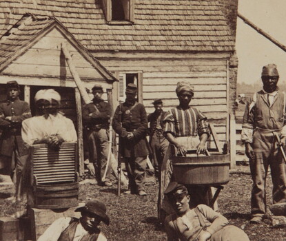 Black men and women laying on the ground and standing (those standing holding tools, such as washboards and basins and an axe) in front of Union soldiers in uniform, themselves in front of a house.