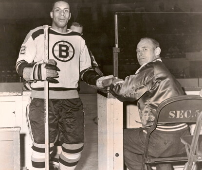 A black-and-white photograph of Willie O'Ree, a Black man, coming off the ice wearing a white Boston Bruins uniform.