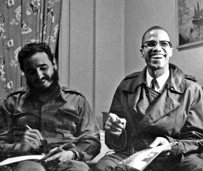 A black-and-white photogrpah of Fidel Castro (a dark-haired, bearded, light-skinned Latino man wearing a dark shirt and pants) and Malcolm X (a light-skinned Black man wearing horn-rimmed glasses, a dark trench coat, and a white shirt and dark tie) smiling while sitting on a bed in a hotel room.