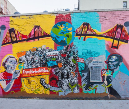 A color photograph of a multicolored mural featuring images of crowds of protesters, Yuri Kochiyama (a Japanese American woman depicted wearing a red shirt and white bandana) speaking into a megaphone, and Malcolm X (a Black man depicted wearing a light-colored suit, blue shirt, and red tie and holding a newspaper). Above them is a depiction of Earth flanked by bridges.