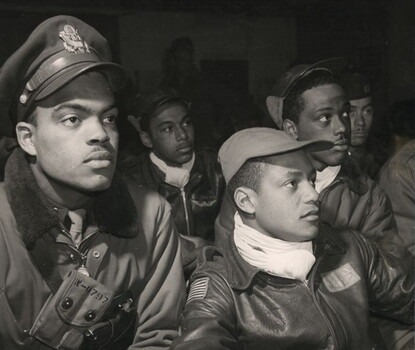 A black-and-white photograph of Black men in WWII aviator uniforms sitting, their attention directed out of frame.