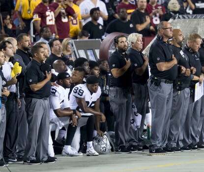 A color photograph of two Black teammates from the Oakland Raiders in the team uniform kneeling on one knee as coaches and staff around them stand with right hands over their hearts.