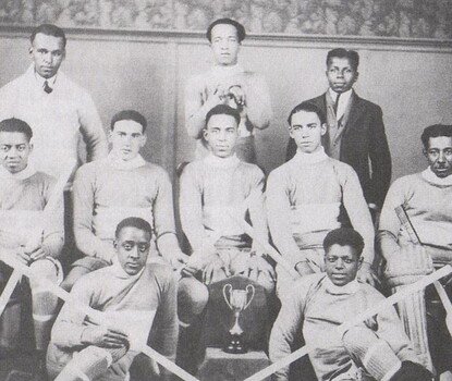 A black-and-white team photo of Black hockey players in light colored uniforms, seated with hockey sticks at their side, surrounding a small trophy.