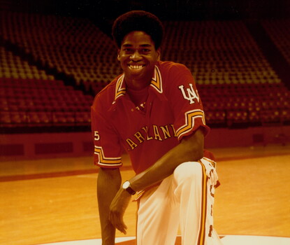 A color photograph of a college-aged Len Elmore kneeling on a basketball court wearing a red Maryland collared shirt, white pants, and white sneakers, his right hand on a basketball and resting his left elbow on his left knee.
