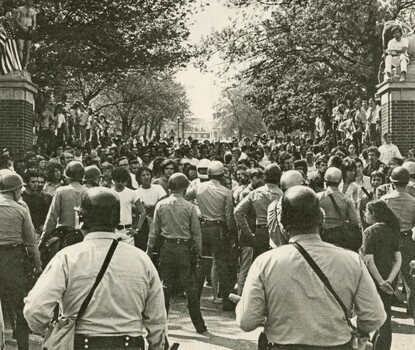 A color photograph of large number of students on a campus pathway, flanked at either end by brick gates. In front of them are campus police officers in uniform, some wearing helmets.