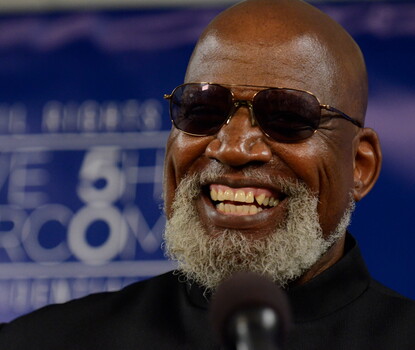 A color photograph of Harry Edwards, a smiling bald Black man with a gray beard, wearing sunglasses and a dark Nehru jacket.