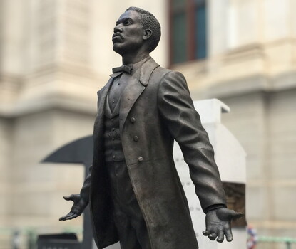 A color photograph of a bronze statue of Octavius V. Catto in a three-piece suit and bowtie, leaning forward, hands open, in the courtyard of Philadelphia City Hall.