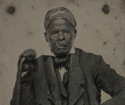 A black-and-white photograph of a Black man wearing a dark turban, jacket, coat, and tie, seated, holding a cane in his right hand.