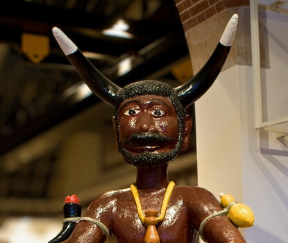 A color photograph of a statue of Papa Legba, who appears as a bearded Black man with black and white horns, a necklace, and gourds and peppers strapped to his arms.