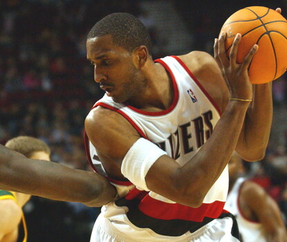 A color photgraph of Shareef Abdur-Rahim, a light-skinned Black man wearing a white Portland Trail Blazers uniform, holds a basketball as Reggie Evans (a dark-skinned Black man in a green and yellow Seattle SuperSonics uniform) defends.